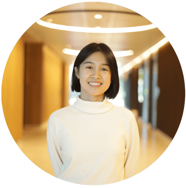 Portrait of a young woman with short black hair, wearing a white turtleneck.  She is smiling with her head gently tilted, hands behind her back, standing in a warmly  lit hallway with a softly blurred background.